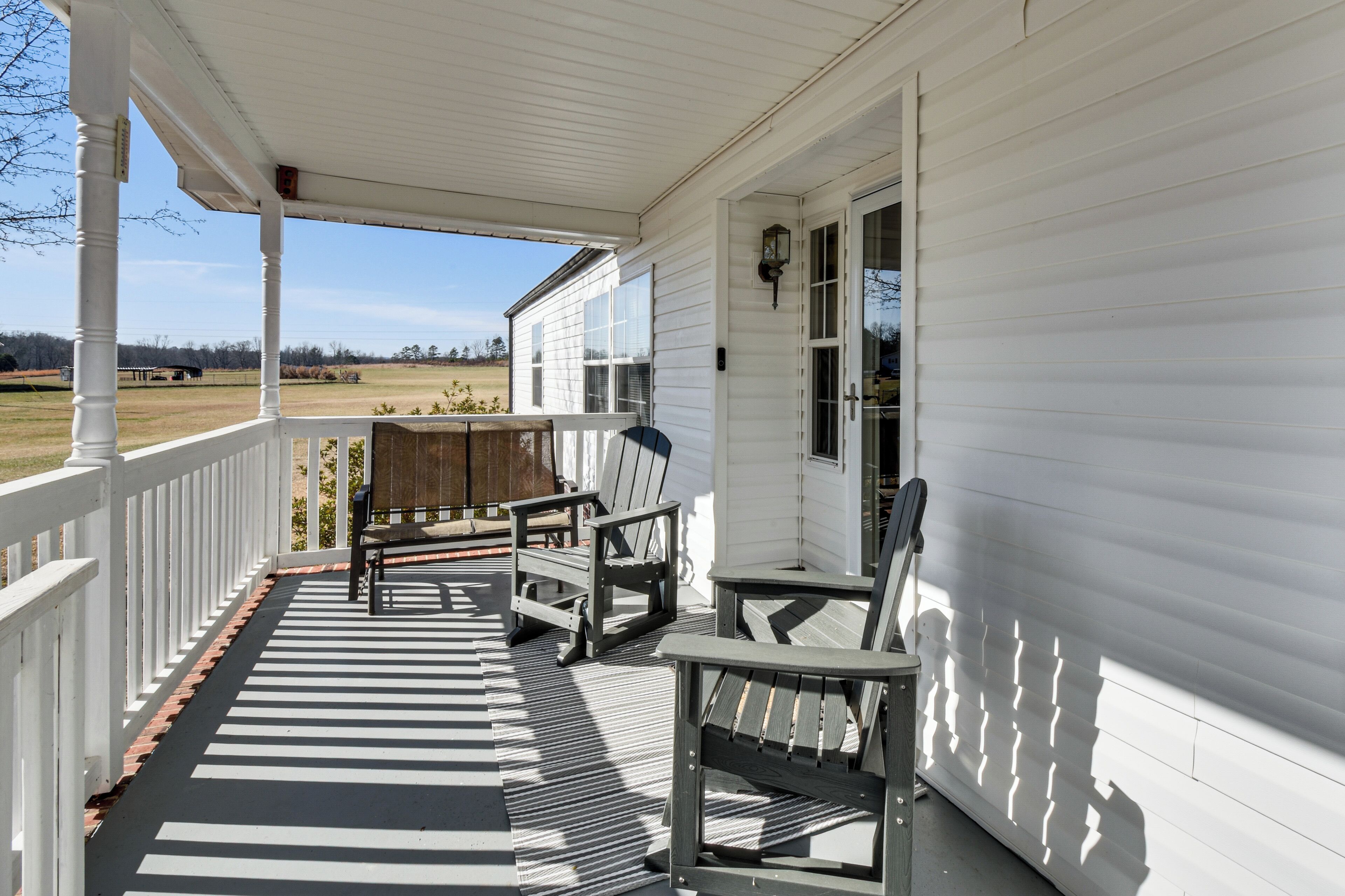A porch with a wooden bench and a chair, a white railing, and a view of a field.