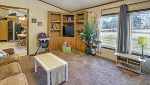 Een woonkamer met een televisie, een groene opbergbox voor speelgoed, een grote potplant en een raam met gordijnen.