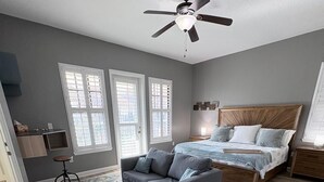 Un dormitorio con cama, sofá gris, un pequeño reposapiés, ventilador de techo y un amplio ventanal con persianas.