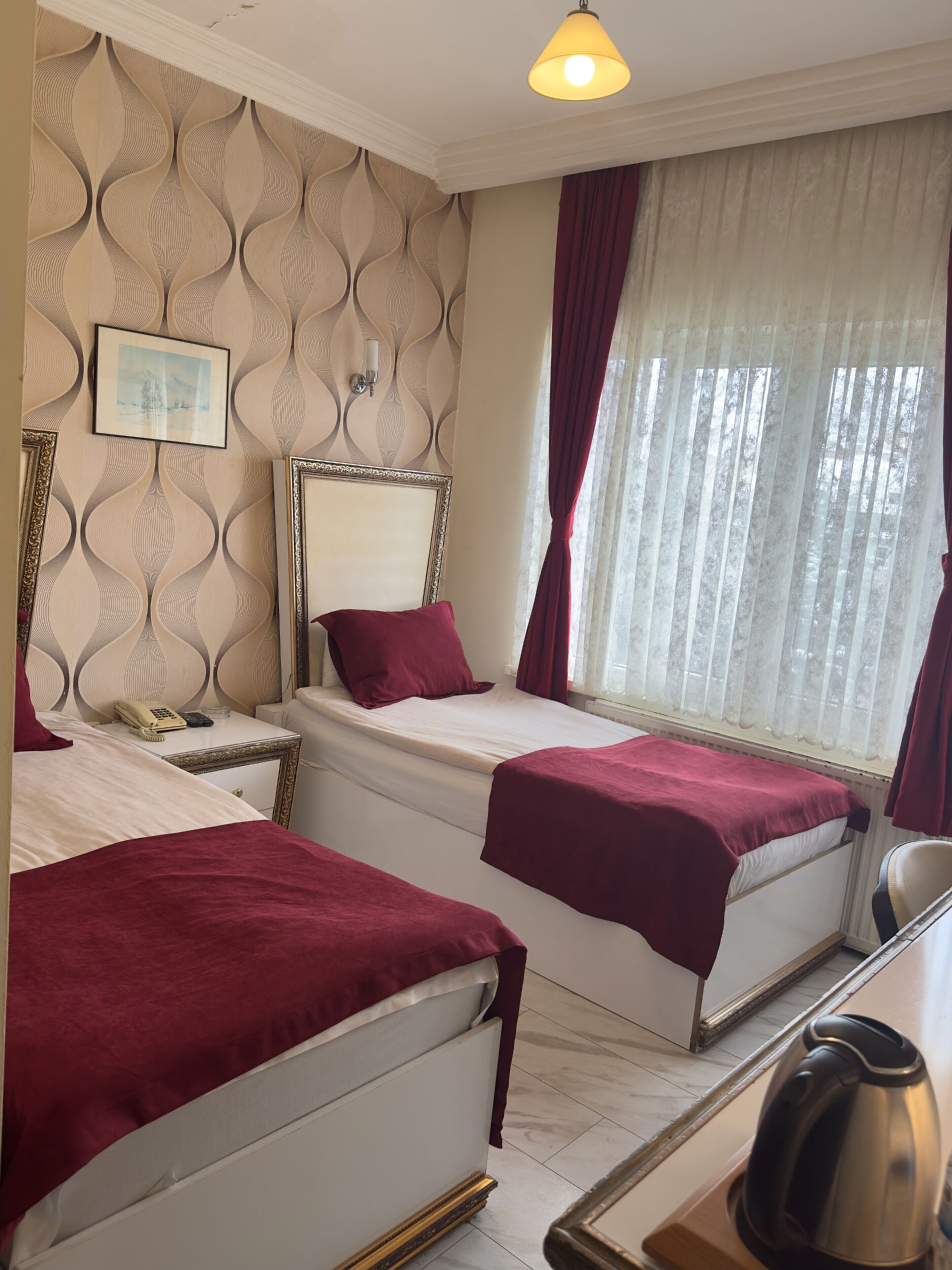 Habitación de hotel con dos camas, una mesita de noche con teléfono, una lámpara y vistas al exterior a través de cortinas translúcidas.