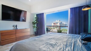 Ein Schlafzimmer mit einem großen Bett, einem an der Wand befestigten Fernseher, einer hölzernen Kommode und einem Balkon mit Blick auf ein Kreuzfahrtschiff.