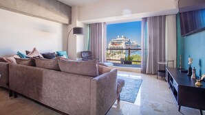Ein modernes Wohnzimmer mit einer großen Couch, einem Flachbildfernseher und Blick auf ein Kreuzfahrtschiff durch große Fenster.