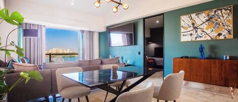 A modern living room with a glass dining table, comfortable seating, a large TV, and a view of the cityscape.