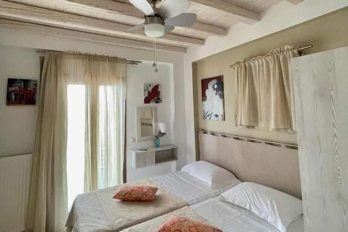 Habitación de hotel con dos camas, ventilador de techo, ventana con cortinas y una mesita de noche pequeña.