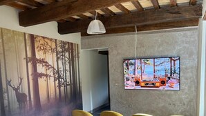 A room with a rustic wooden ceiling, a large mural of a forest scene, and a television mounted on the wall. There are six chairs with wooden legs and colorful upholstery arranged in front of the TV. - Apt renovated in farmohouse with panoramic pool (Città di Castello)