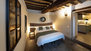 A bedroom with a wooden ceiling, a bed with a white bedspread, a round mirror above the bed, and a small kitchen area to the right. - Apt renovated in farmohouse with panoramic pool (Città di Castello)