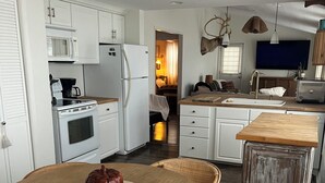 Une cuisine moderne avec des meubles blancs, un coin repas avec des chaises en osier et une table en bois, et un salon chaleureux doté d’une cheminée.