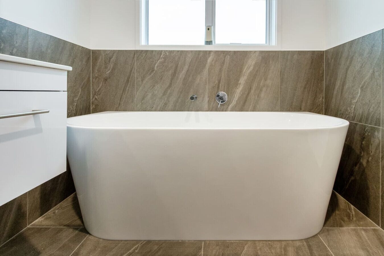 A modern bathroom with a freestanding bathtub, a white cabinet, and a window above the tub.