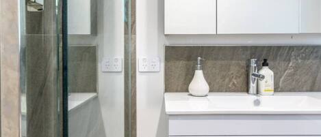 Un baño moderno con cabina de ducha de vidrio, tocador blanco con encimera de mármol y calentador de pared.