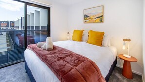 A hotel room with a large bed, a view through a sliding glass door, a painting on the wall, and a bedside table with a lamp. - Central City | Walk to Stadium, Dining & Shopping (Christchurch)