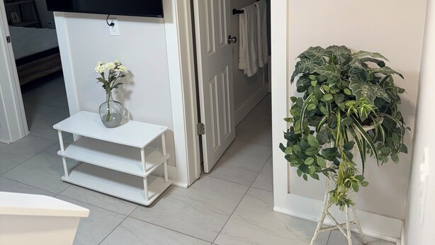 Une pièce dotée d’un téléviseur à écran plat fixé au mur, d’une petite table blanche ornée d’un vase de fleurs et d’une plante en pot posée sur un support.
