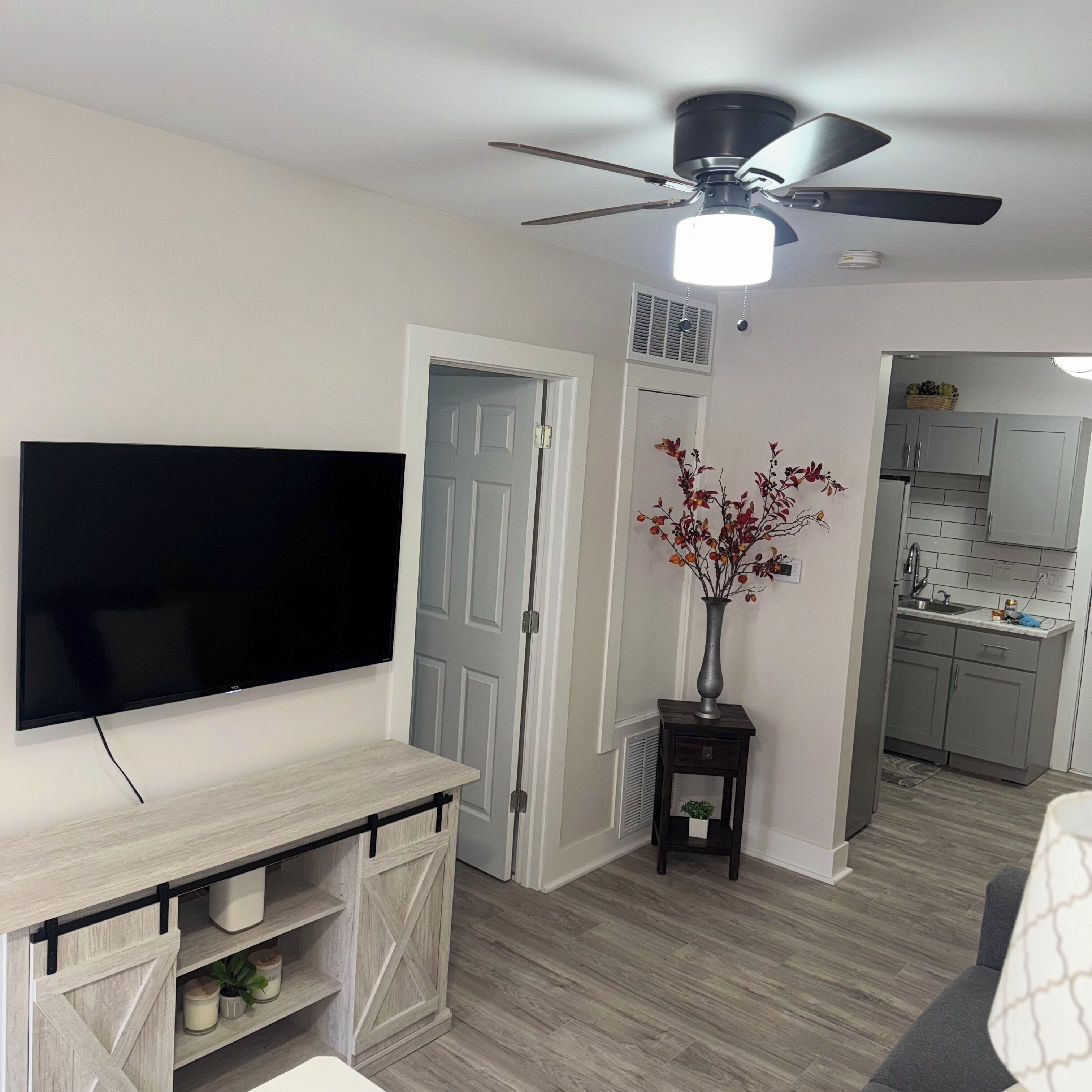 Una sala de estar con televisor de pantalla plana, ventilador de techo, un florero con flores y una cocina visible a través de una puerta abierta.