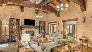 A rustic living room with stone walls, wooden beams, a fireplace, and a large flat-screen TV. - Villa Di Gioia by AvantStay | Private Wine-Country Estate + Vineyard Views • Near Top Temecula Wineries (Temecula)