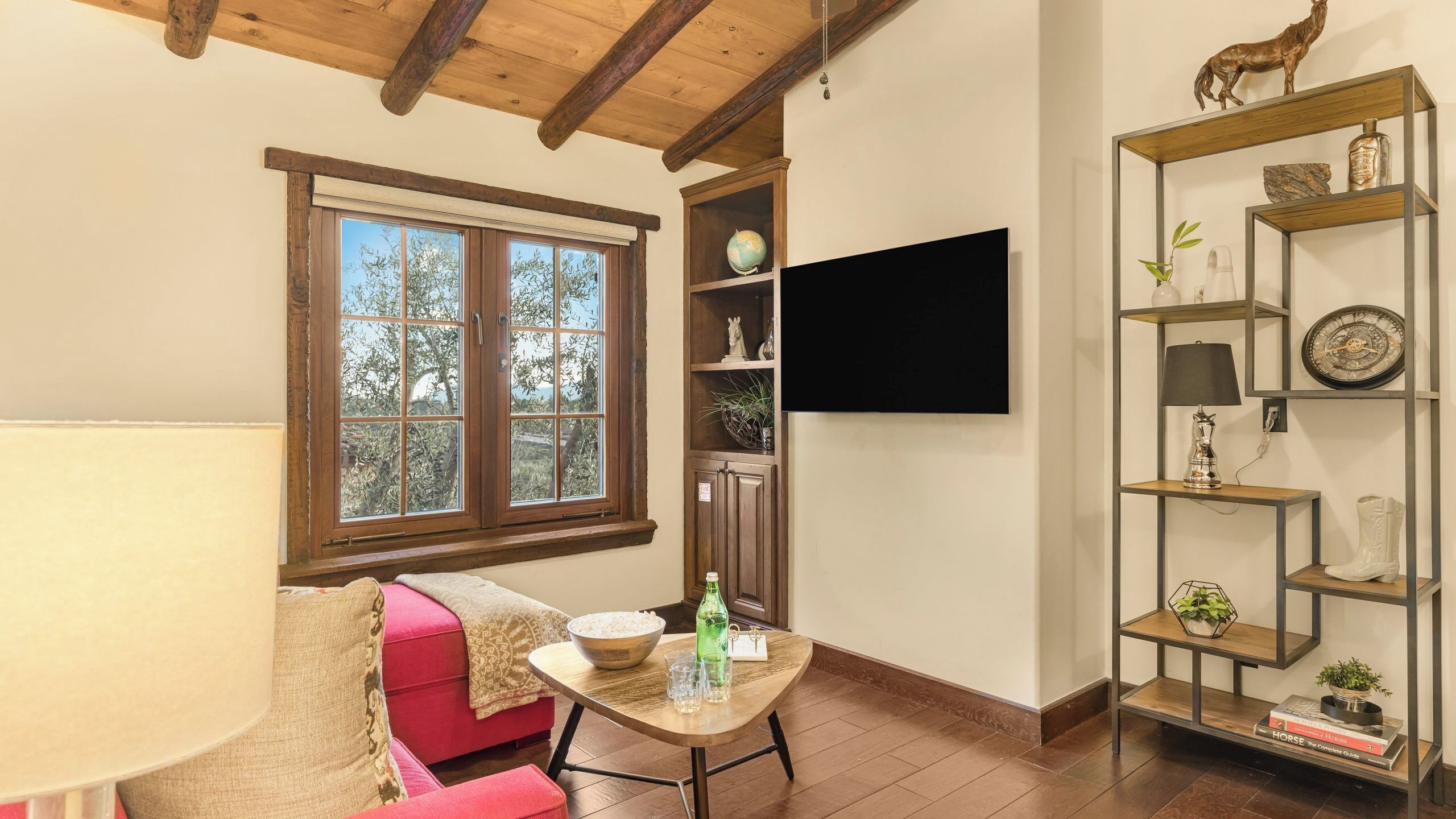 A cozy living room with a wooden ceiling, a large window, a pink sofa, a small table with drinks, and a bookshelf with decorative items.