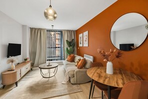 Egy modern nappali, melyben található egy kanapé, egy kerek, fából készült asztal, egy televízió és egy nagy ablak, amelyen függöny van.