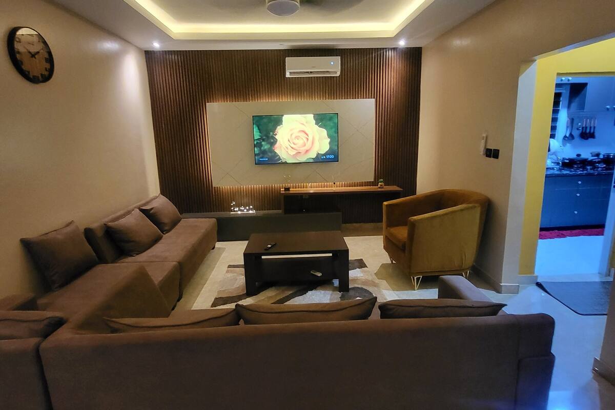 A modern living room with a sectional sofa, a coffee table, and a flat-screen TV mounted on the wall.