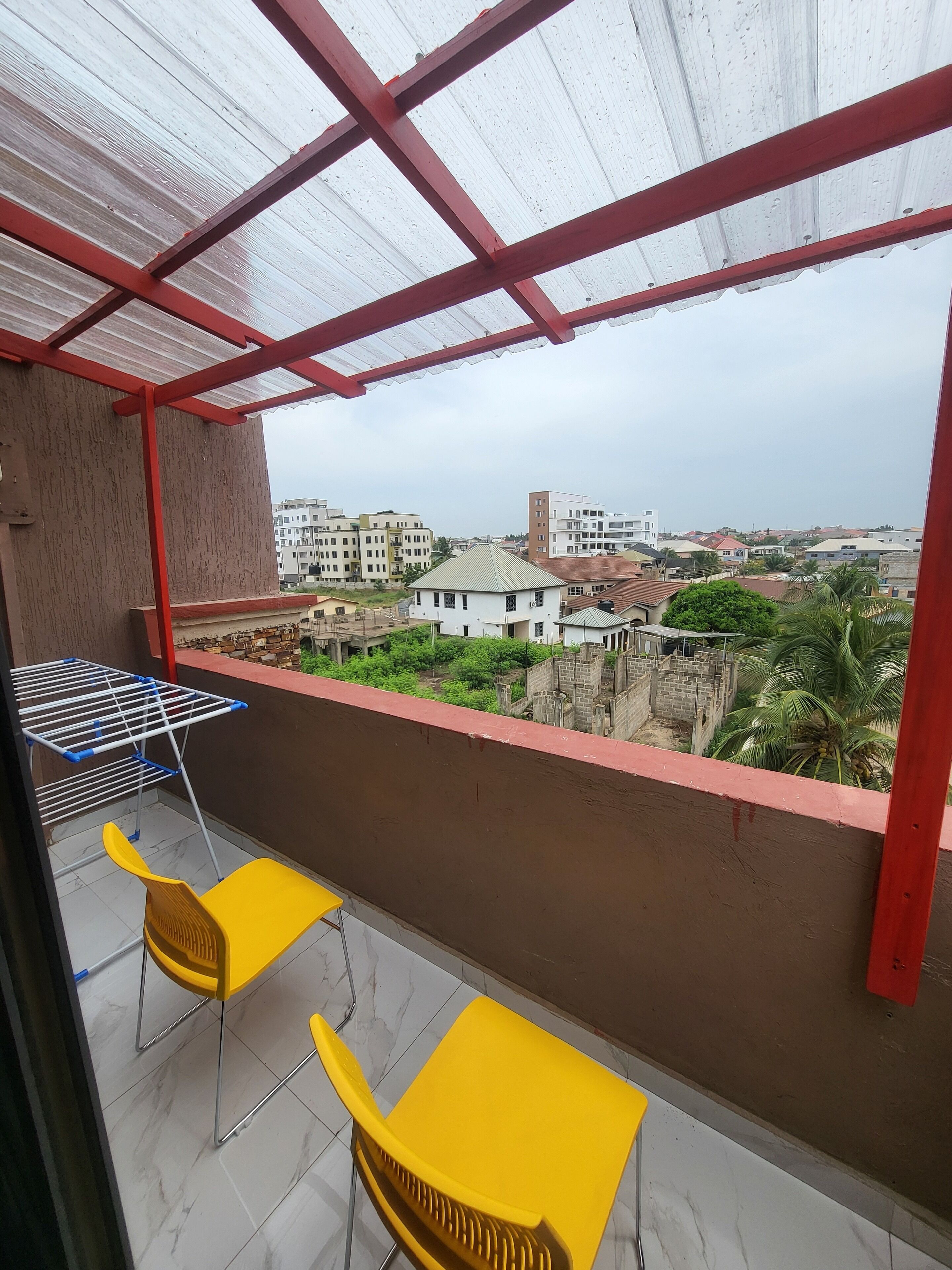 Un balcón con estructura de metal rojo, suelo de baldosas blancas y vistas a edificios y vegetación.