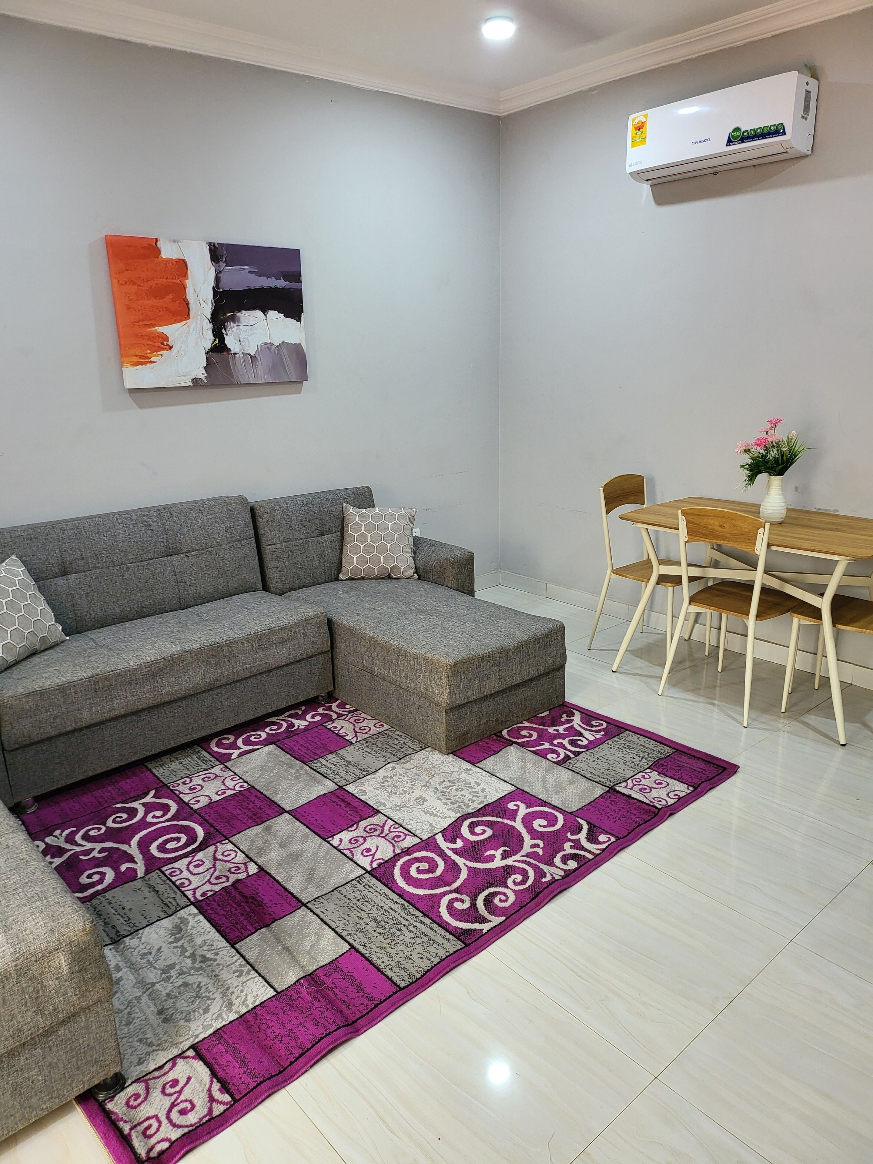 A living room with a grey sectional sofa, a wooden dining table with chairs, a purple and grey patterned rug, and a wall-mounted air conditioner.