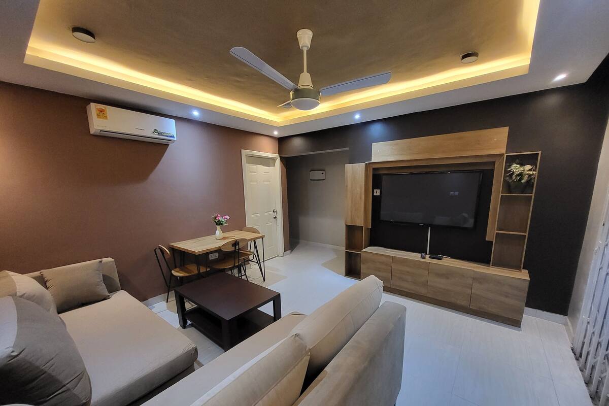 A modern living room with a sofa, a dining table with chairs, a TV mounted on a wooden wall, and a ceiling fan.