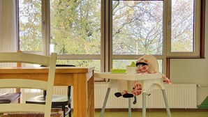 A room with a dining table and chairs, a high chair, and large windows with a view of trees. - Amsterdam Farm Lodge (Amsterdam)