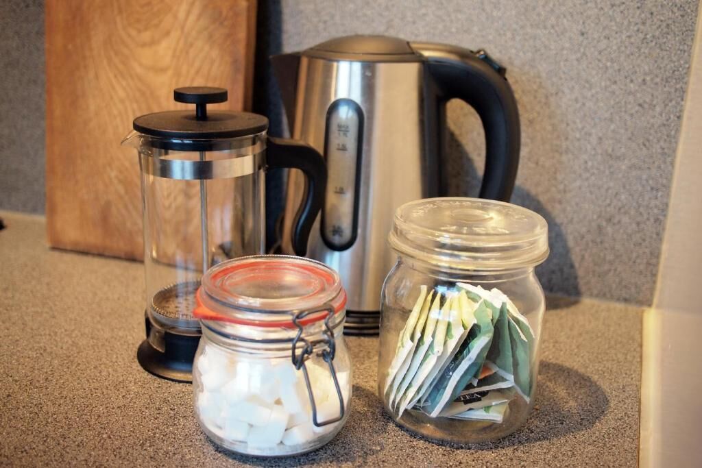 Eine Küchenarbeitsplatte mit einer Kaffeepresse, einem Edelstahl-Wasserkocher und zwei Glasbehältern mit weißen Würfeln und grünen Papierstücken.