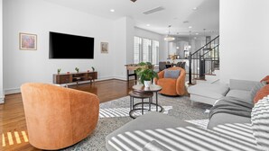 Tilava olohuone, jossa on litteänäyttöinen televisio, oranssit ja harmaat sohvat, puinen sohvapöytä ja portaat, jotka johtavat ylemmälle tasolle.