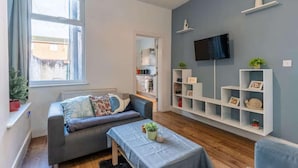 Ett modernt vardagsrum med en grå soffa, ett soffbord och en tv som är monterad på väggen. I bakgrunden syns vita hyllsystem med dekorativa föremål och ett litet kök.
