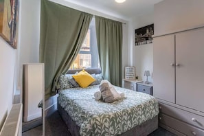A bedroom with a bed, green curtains, a bedside table with a lamp, a wardrobe, and a wall with a framed picture. - The Kensington House (Liverpool)