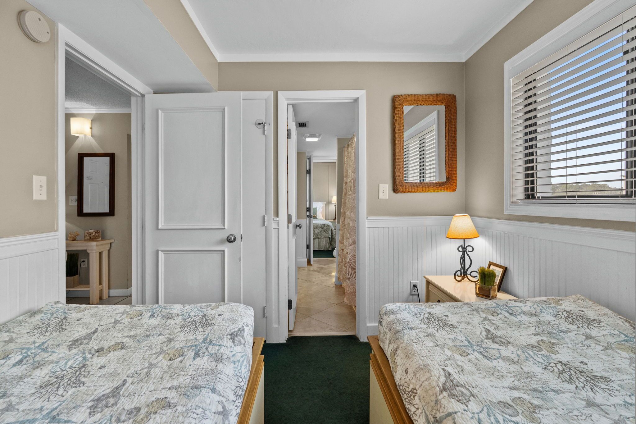 A hotel room with two beds, a mirror, a window with blinds, a lamp, and a door leading to another room.