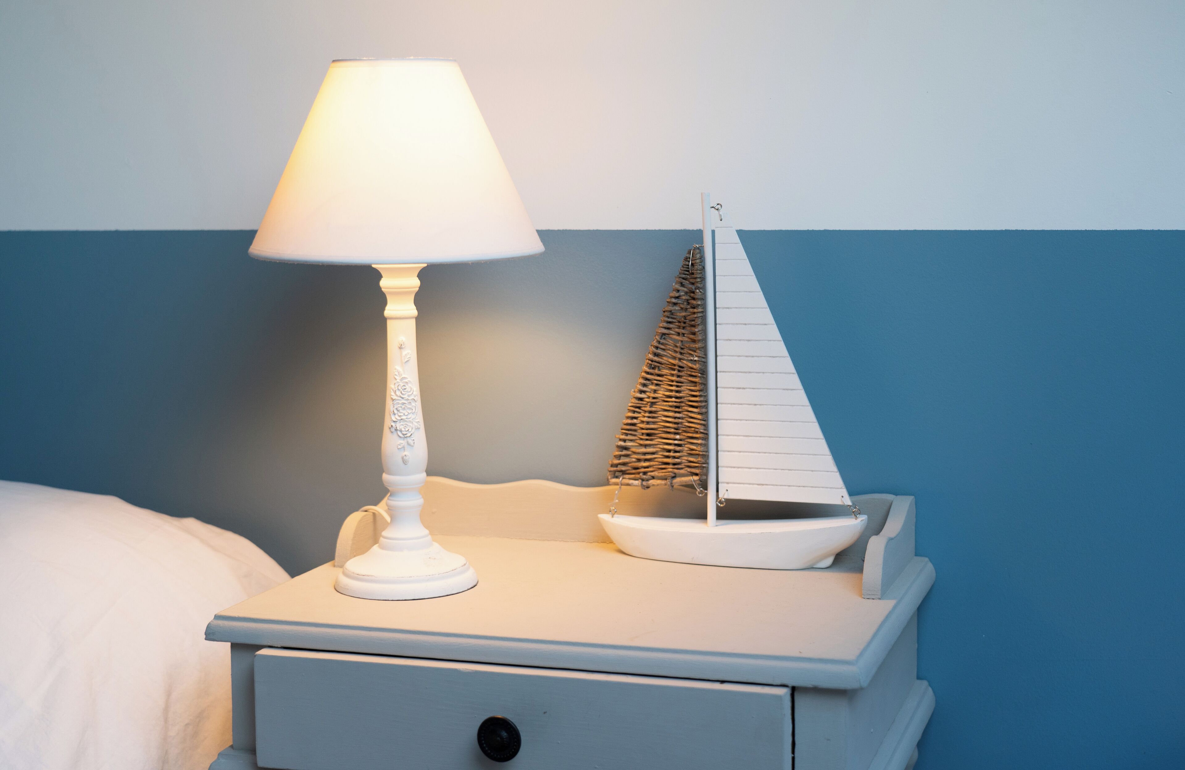 Une table de chevet avec une lampe blanche et un petit modèle de voilier blanc.