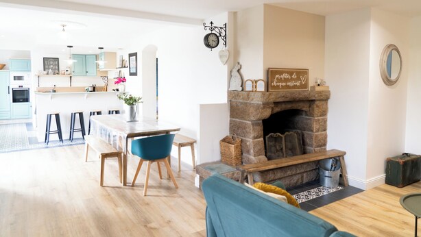 A modern living room with a stone fireplace, a dining area with a long table and blue chairs, and a kitchen in the background.