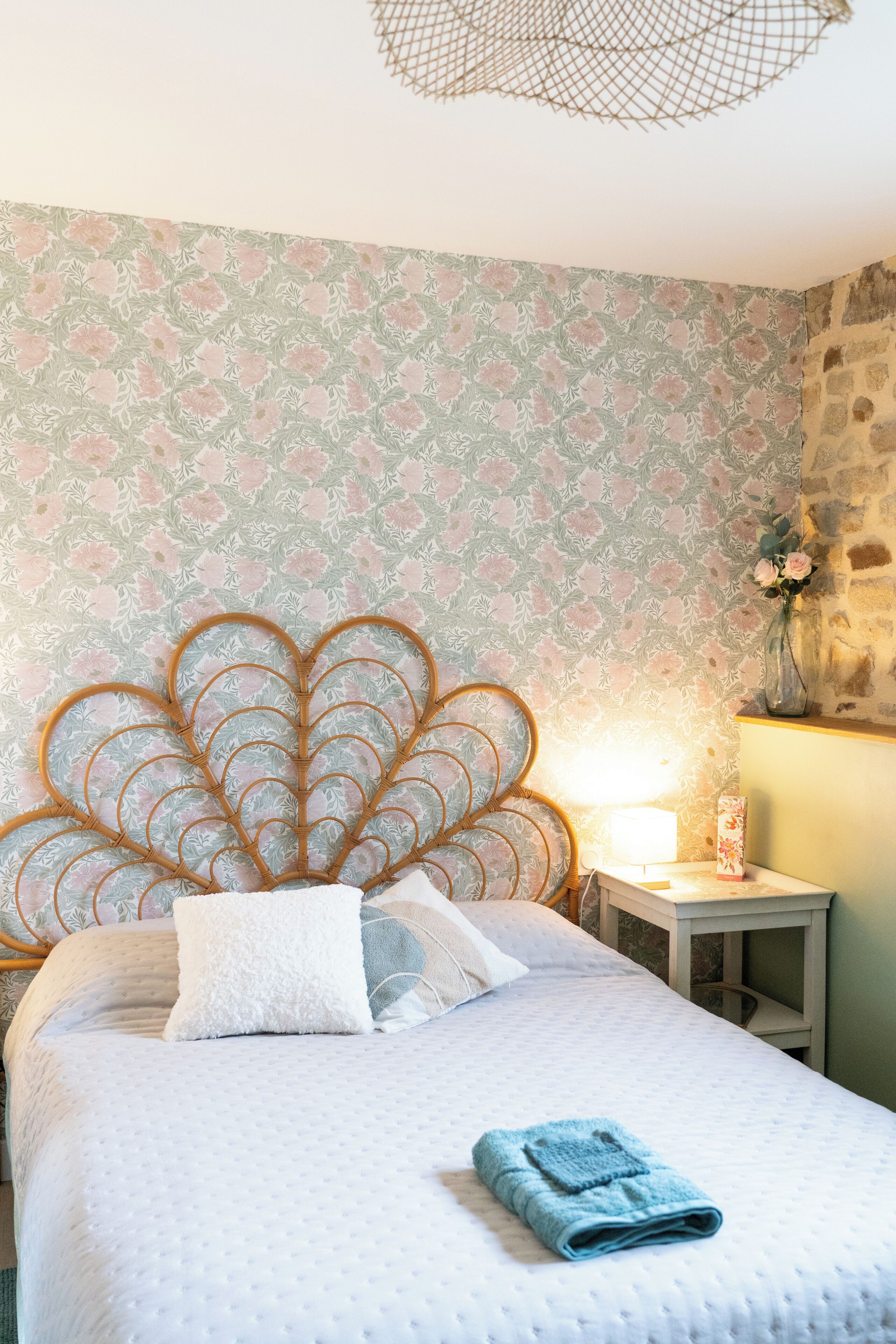 A neatly made bed with a white duvet and a blue towel, a wooden headboard with a scalloped design, a small bedside table with a lamp, and a patterned wallpaper.