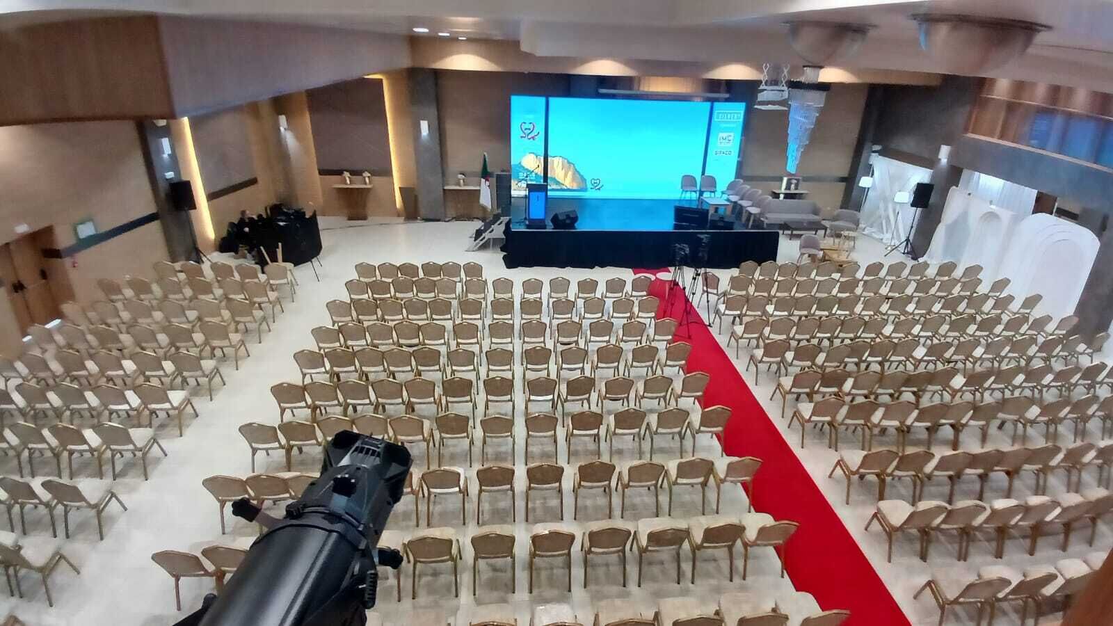 Un salón de conferencias con filas de sillas, un escenario con una pantalla y una alfombra roja.