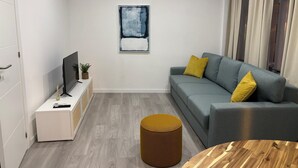 Ett modernt vardagsrum med en grå soffa, gula kuddar, en tv på ett vitt skåp, en tavla på väggen och ett runt träbord.