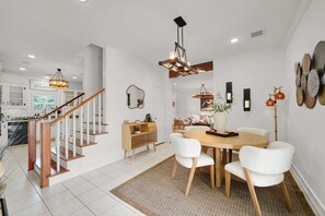 Eine moderne Küche mit weißen Schränken, einer Holztreppe, einem Essbereich mit rundem Tisch und weißen Stühlen sowie einem Wohnzimmer mit Sofa und Couchtisch.