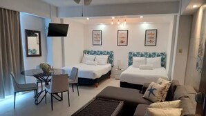 Ein Hotelzimmer mit zwei Betten, einem Esstisch mit Stühlen, einer Couch, einem kleinen Tisch und einem an der Wand montierten Fernseher.