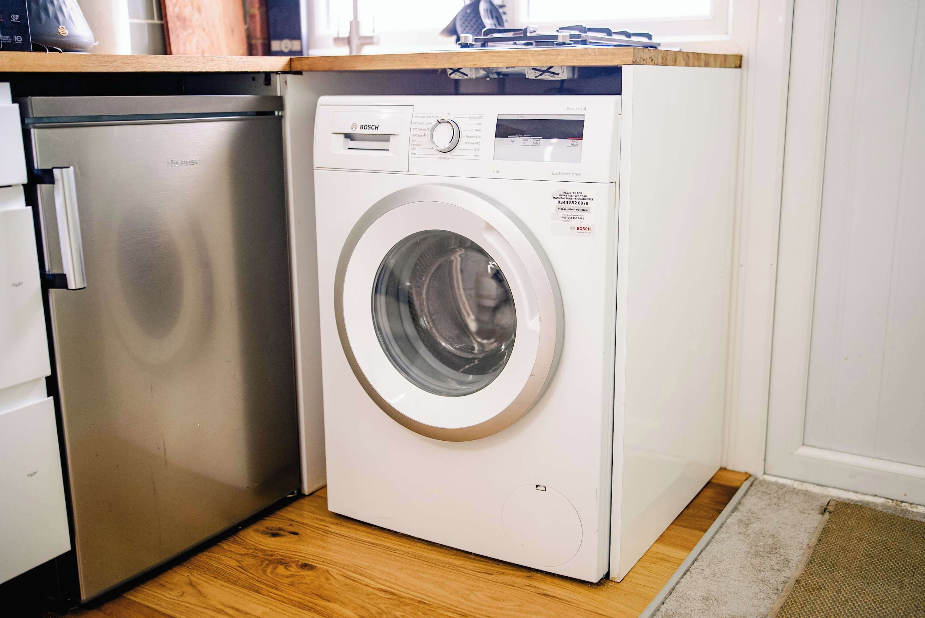 A compact, white Bosch washing machine in a corner of a room with wooden flooring and a small window.