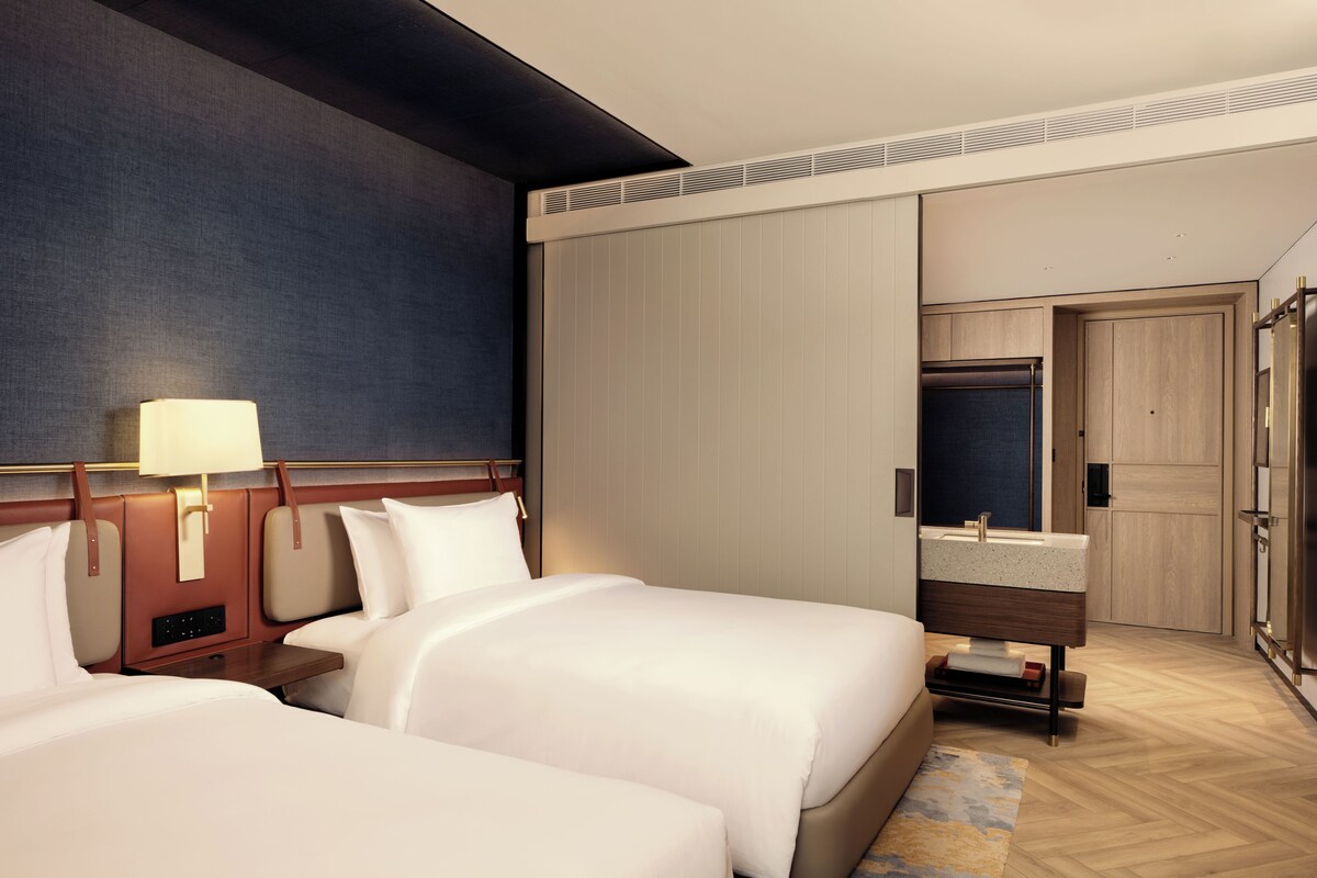Une chambre d’hôtel avec deux lits, une salle de bain avec un lavabo et un miroir, et une armoire.