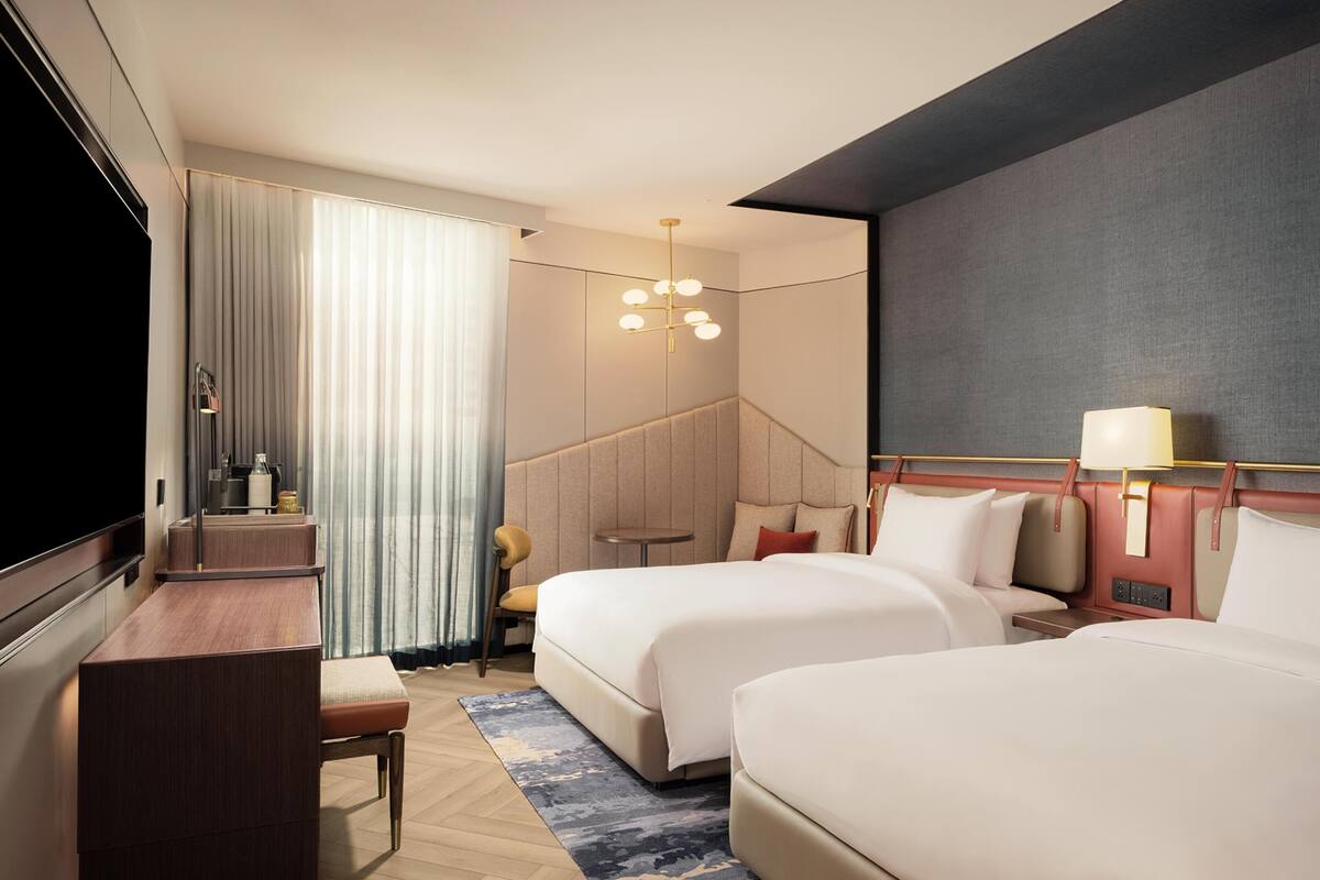 Habitación de hotel con dos camas, un escritorio, una silla, un televisor, una lámpara y una ventana con cortinas.