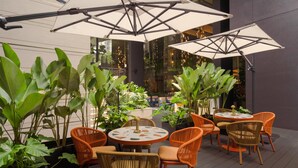 An outdoor patio area with round tables and orange wicker chairs, surrounded by large potted plants. - Canopy By Hilton Bangkok Sukhumvit (Bangkok)