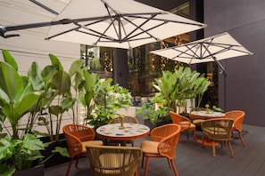 Ein Terrassenbereich im Freien mit runden Tischen und orangefarbenen Korbstühlen, umgeben von großen Kübelpflanzen.