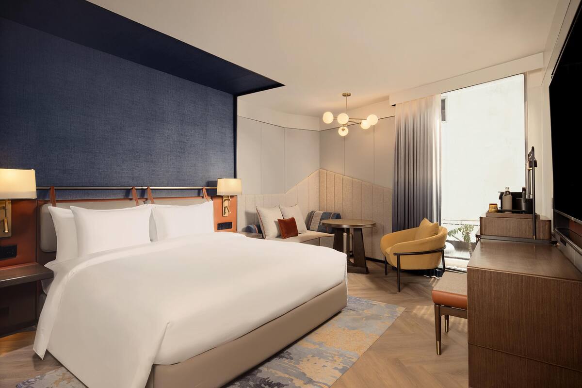 Habitación de hotel con una cama grande, escritorio, silla, televisor y balcón con cortinas.