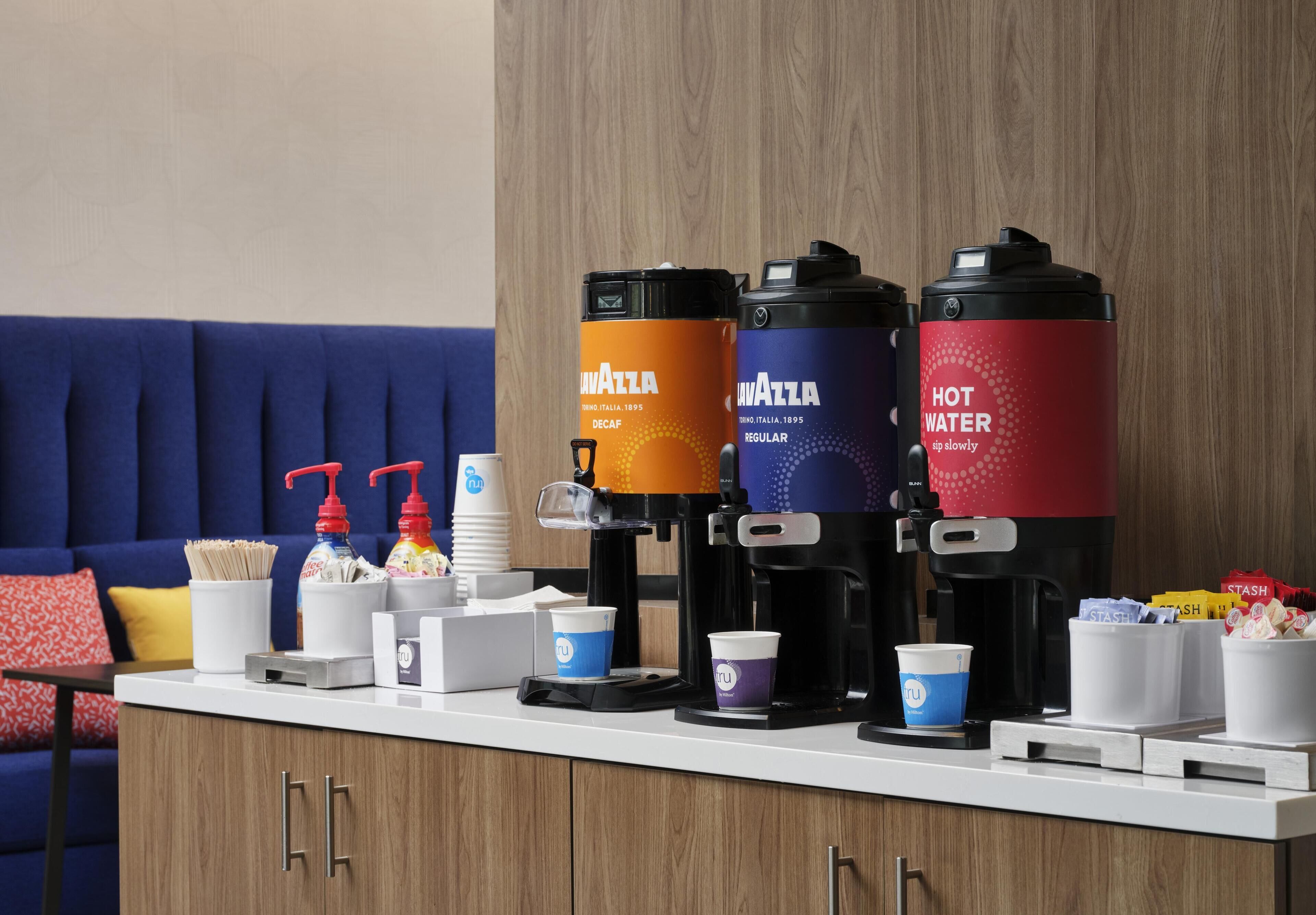 A coffee station with three dispensers labeled 'Decaf,' 'Regular,' and 'Hot Water,' and a variety of disposable cups.