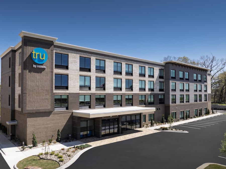 A modern hotel building with a prominent logo, multiple windows, and a parking lot.