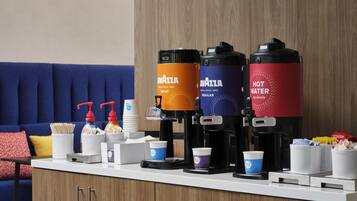 Una camera d'albergo con un angolo caffè dotato di tre erogatori, un divano e un tavolo con tazze e condimenti.