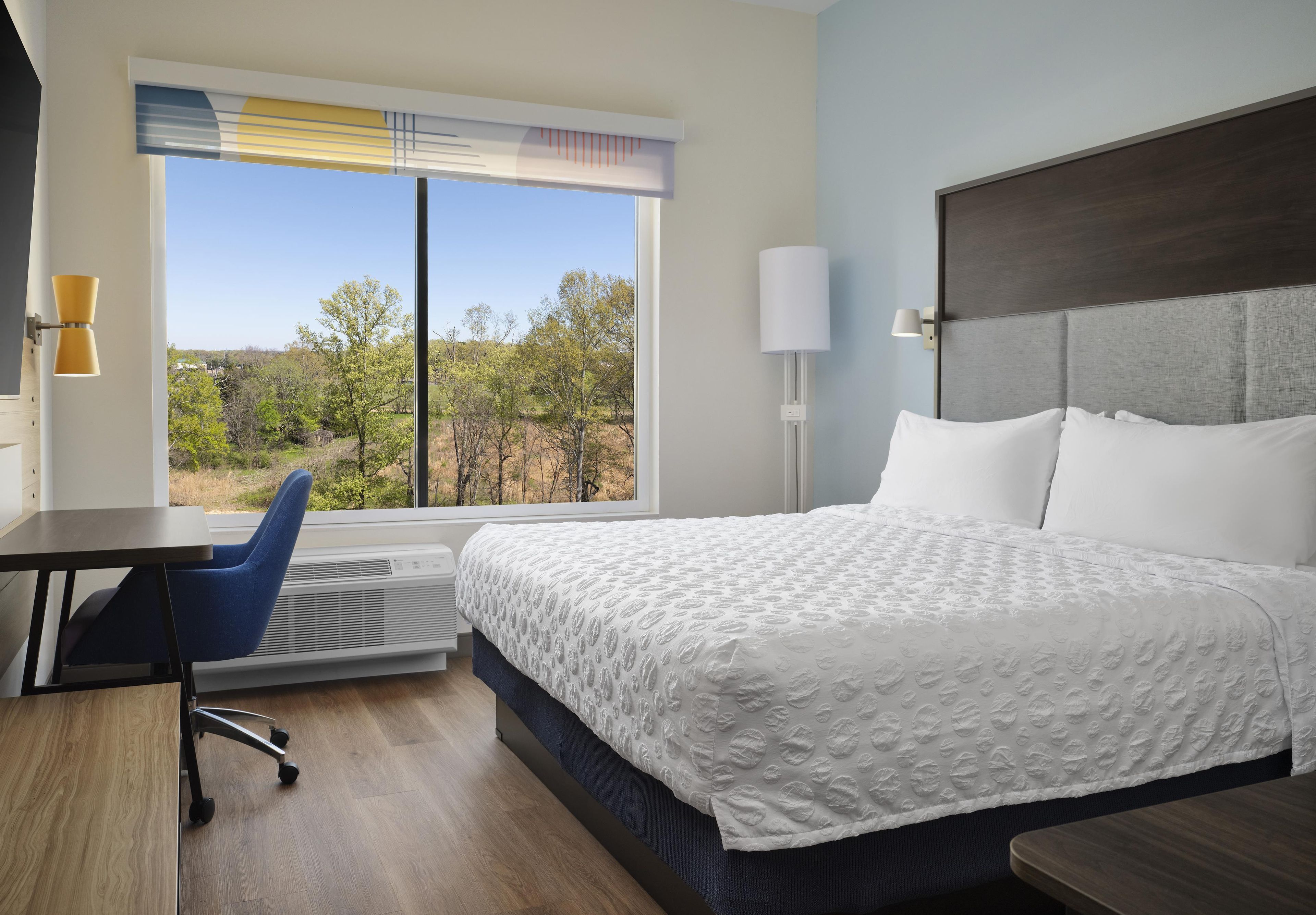 Una habitación de hotel con cama, escritorio, silla y un amplio ventanal con vista a árboles y cielo despejado.