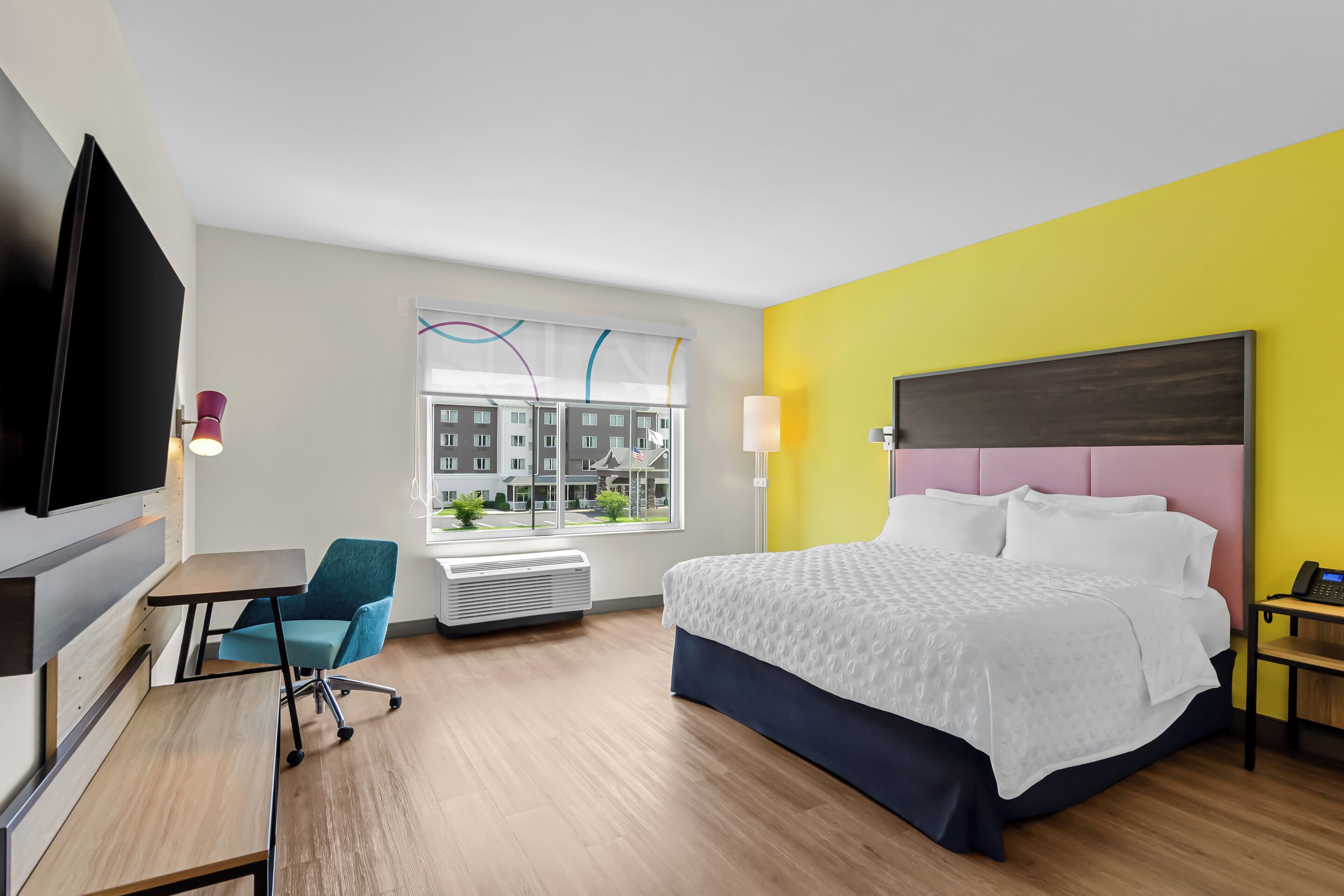 A hotel room with a bed, a desk, a chair, a TV, and a window with a view of buildings.