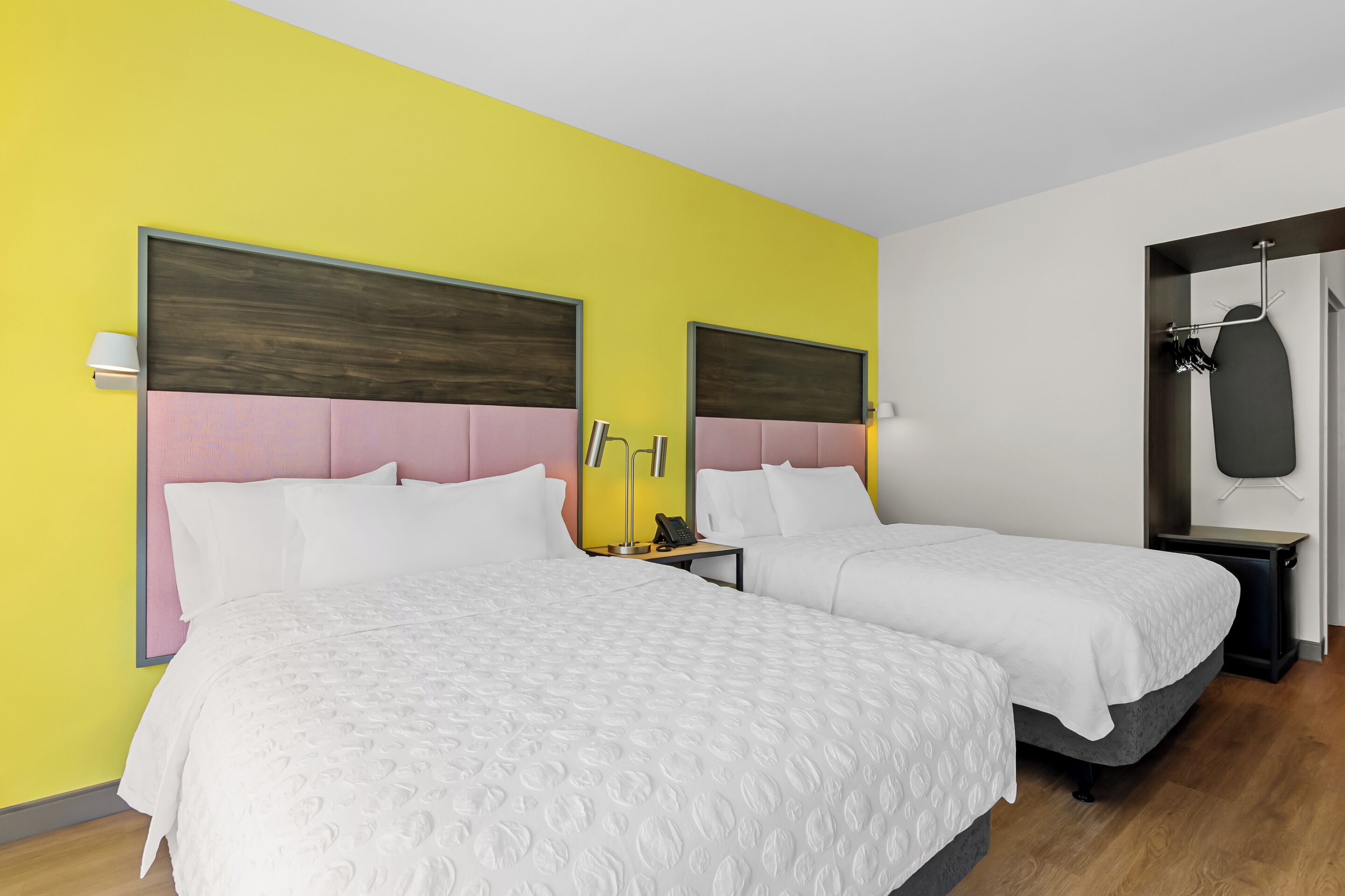 A hotel room with two beds, a wooden headboard, a bedside table with a lamp, and a coat rack.