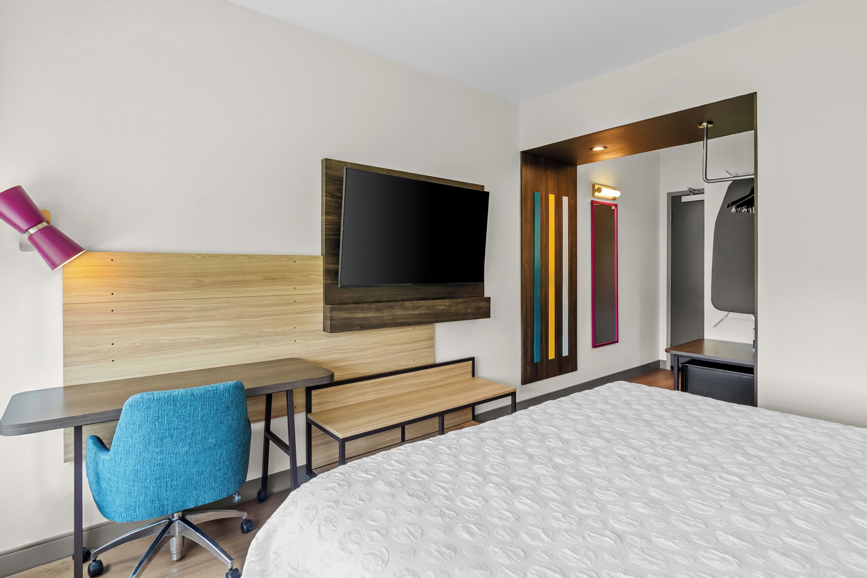 A hotel room with a bed, a desk with a chair, a television mounted on the wall, and a mirror with colorful vertical stripes.
