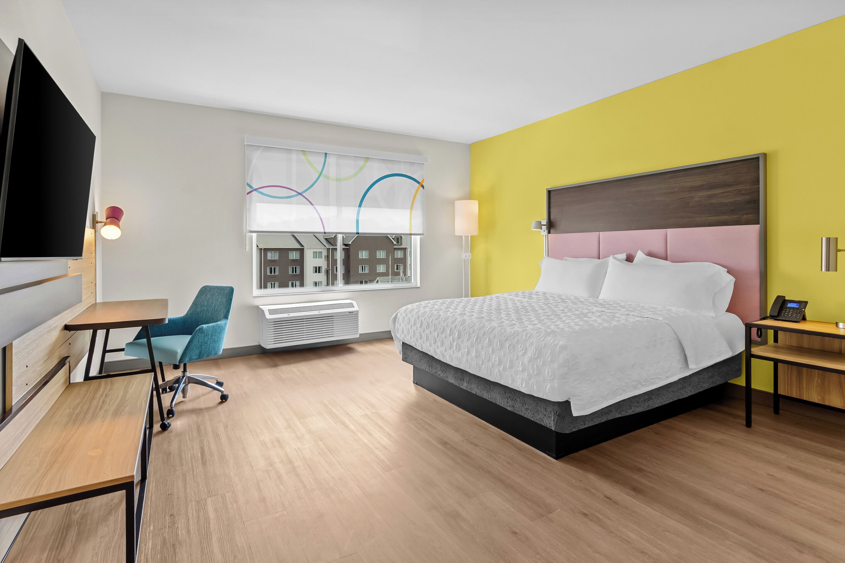 A hotel room with a bed, desk, chair, and a window with a view of buildings.